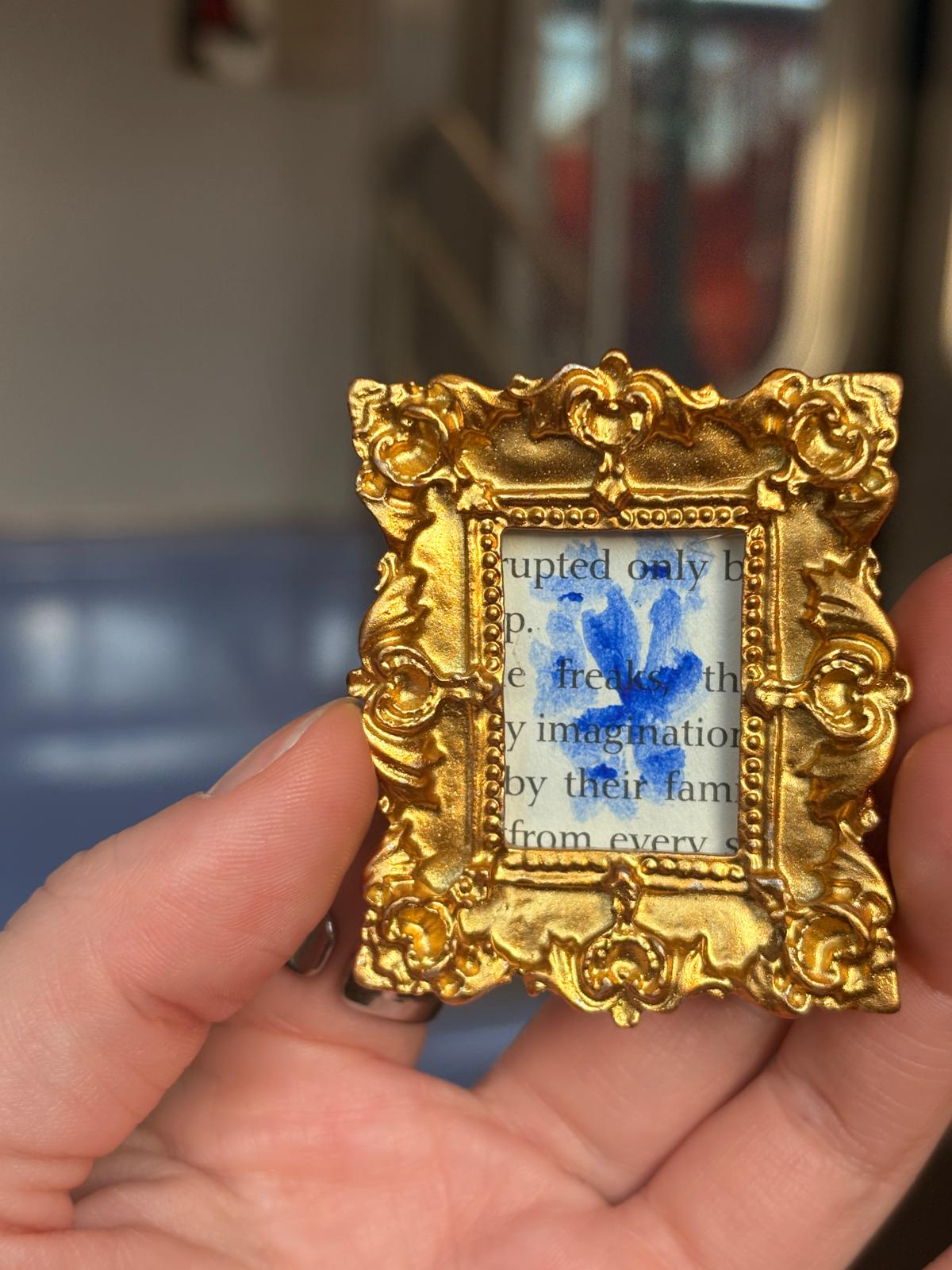 Blue painting on a book page in a tiny gold frame